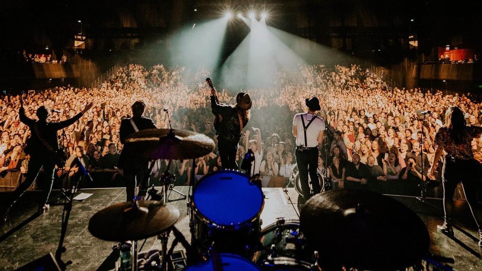 Band playing a sold-out show, shot from behind the drum kit — the kind of tour photo Woodstock picks for the content calendar